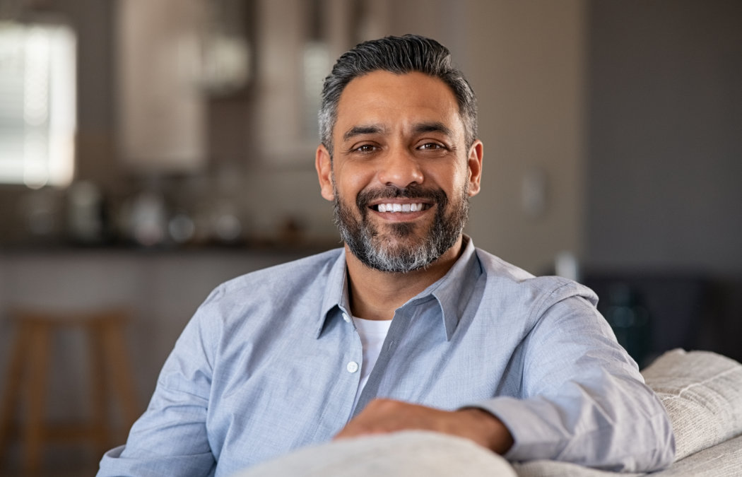 Smiling man with short hair and beard, wearing a light blue shirt, sitting on a sofa in a cozy living room., 