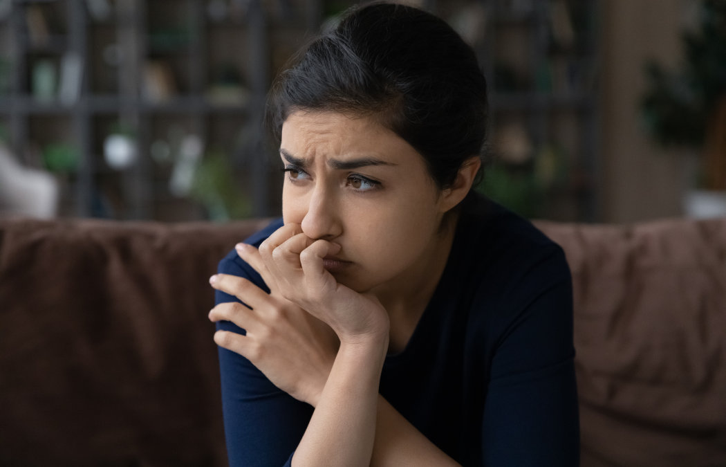 A person with dark hair looks concerned, sitting on a brown couch with their hand covering their mouth. Background shows a blurry bookshelf., 