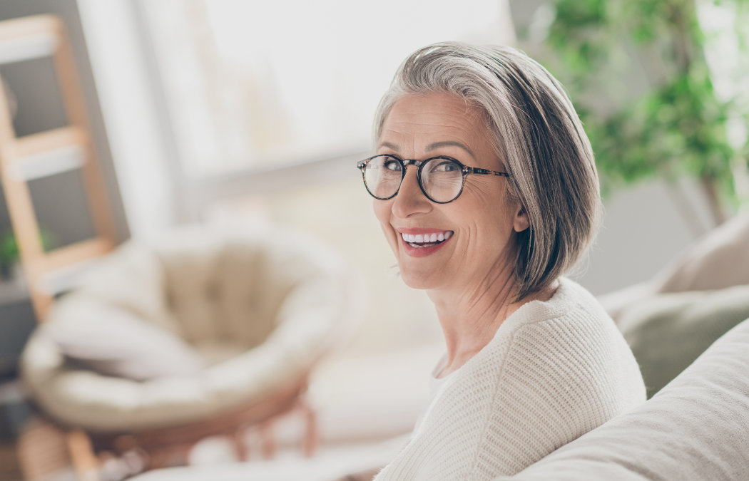An older woman with gray hair and glasses sits on a couch, smiling at the camera. She is wearing a white sweater. The background is softly lit with some plants visible., 