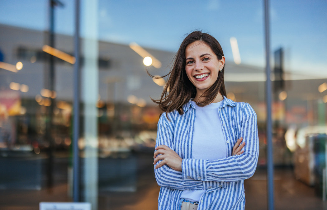 A woman with long brown hair smiles with arms crossed, standing in front of a glass building entrance. She is wearing a white shirt and a blue-and-white striped shirt., 