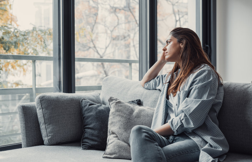 Woman sitting on a couch, looking pensively out a large window. She rests her chin on her hand, with trees visible outside., 