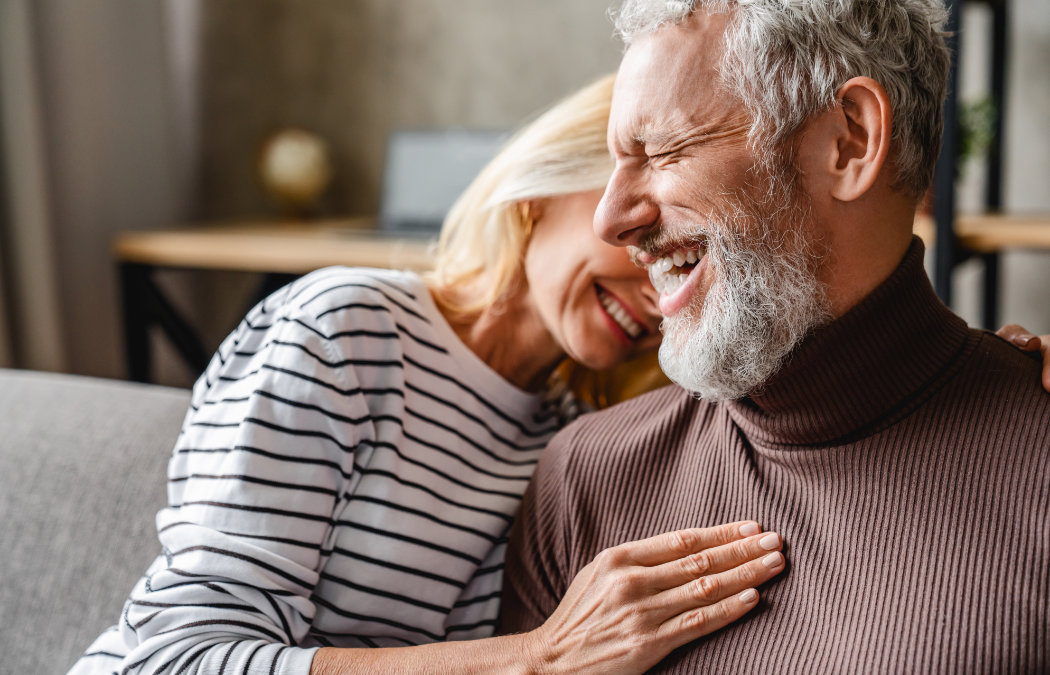 An older woman and man sit close together, smiling and laughing. The woman wears a striped shirt; the man has a gray beard and wears a brown turtleneck sweater., 