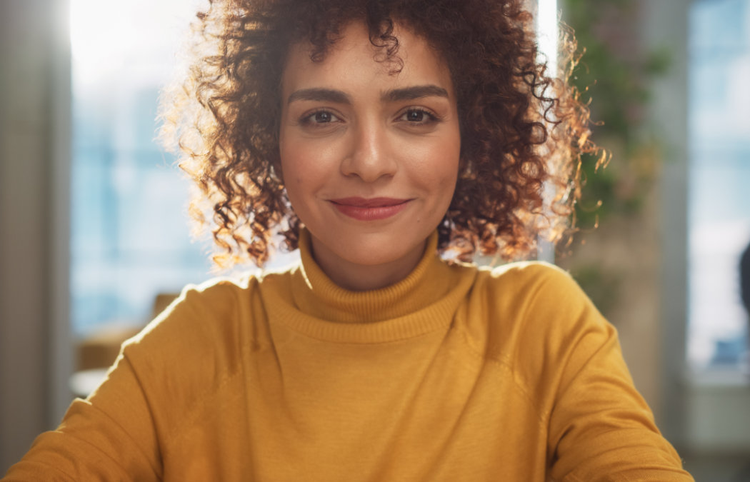 A person with curly hair and a yellow turtleneck sweater smiles while sitting indoors with sunlight in the background., 