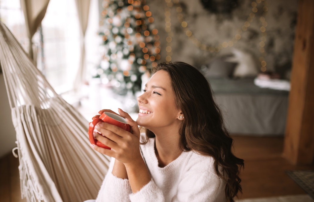 Smiling dark-haired girl dressed in beige sweater and pants holds a red cup sitting in a hammock in a cozy decorated room with a New Year tree, 
