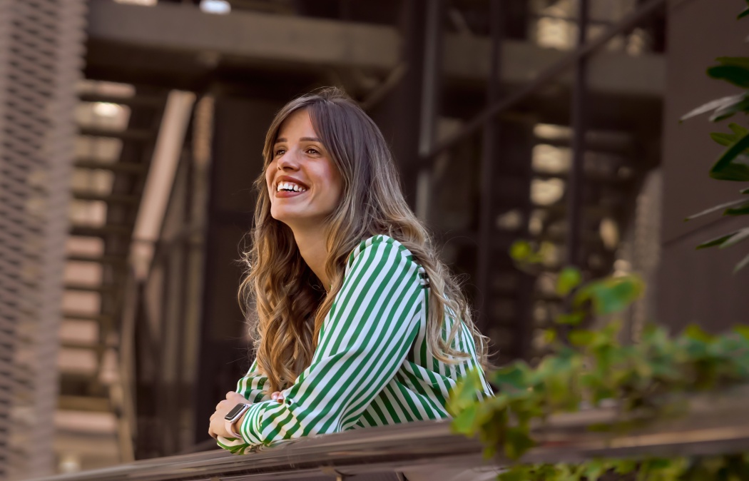Smiling Woman Leaning on Railing in Green Striped Shirt Outdoors contemplating. Portrait of a confident young businesswoman in front of the modern office building., 