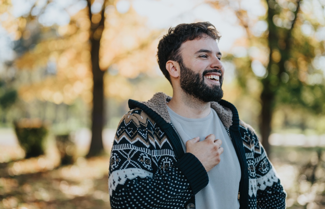 A cheerful man in a warm, patterned sweater stands amidst autumn trees, laughing freely. The sunlit background and fallen leaves capture the essence of an enjoyable, relaxed outdoor moment., 